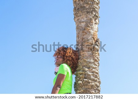 Afro-African girl, relaxed leaning on a palm tree with the sky in the background, with copy space.