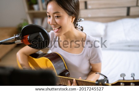 Asian woman playing guitar and recording vocals in her bedroom. She's live. Royalty-Free Stock Photo #2017245527