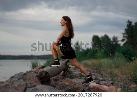Woman in black sport suit is doing a yoga pose (asana), working out outside near beautiful river and stones