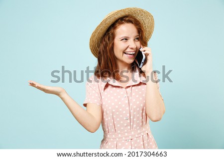 Smiling young redhead curly woman in casual pink dress straw hat hold in hand talk speak on mobile cell phone conducting pleasant conversation isolated on pastel blue color background studio portrait