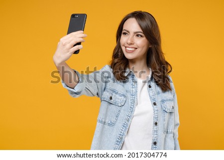 Young smiling happy friendly fun woman 20s wearing stylish denim shirt white t-shirt doing selfie shot on mobile cell phone talking by video call isolated on yellow color background studio portrait.