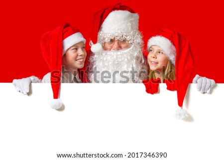 satna claus with funny children in red christmas gnome hats peeking out from behind a blank white banner, mockup and copy space