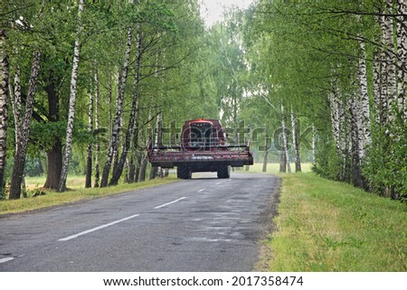 Harvester combine drive on beautiful rustic asphalt road between the Birch trees alley in the village at summer day - harvesting, agriculture, farming in East Europe