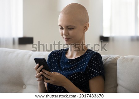 Relaxed young bald after chemotherapy woman sitting on couch, enjoying holding cellphone, web surfing information about cancer treatment, communicating distantly with doctor or using funny apps.