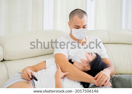 Happy young couple watching television together at home while wearing face mask during self isolation in the living room