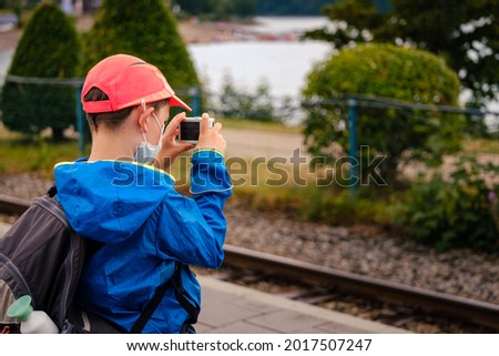 young caucasian boy with face mask wearing orange hat and backpack taking a picture using a digital camera
