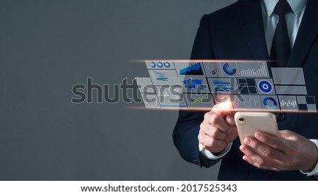 An Asian man in a suit with his left hand holding a mobile phone. His right hand pointing at a hologram showing business icons and stock graph. Black background blur