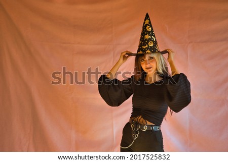 Portrait of young Hispanic woman dressed as a witch holding her hat with both hands on pink background for Halloween.