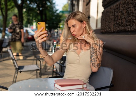 A blonde with a tattoo on her arm sits in a summer cafe and emotionally talks on the phone via video communication. Blogger concept