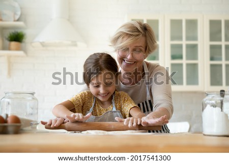 Happy mature grandmother teaching adorable little granddaughter to rolling dough, smiling senior woman in glasses with preschool girl cooking homemade cookies together, family enjoying leisure time Royalty-Free Stock Photo #2017541300