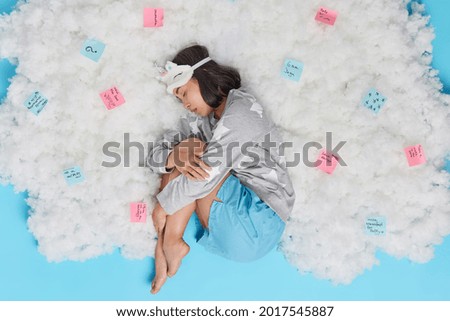 Overhead shot of brunette woman enjoys sleeping wears sleepamsk and pajama falls asleep poses on white cloud with stickers around makes list to do for next day. Relaxation time. Ideas during sleep