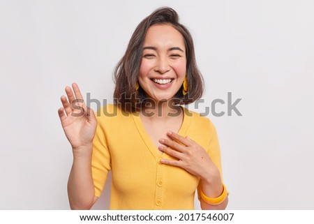 Happy dark haired pretty Asian woman with positive expression laughs joyfully keeps hand raised smiles broadly wears yellow jumper earrings hears something funny isolated over white background. Royalty-Free Stock Photo #2017546007