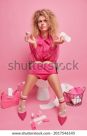Upset curly haired young woman holds sanitary napkin and cotton tampon compares two variants of women hygiene wears fashionable outfit prepares for formal meeting poses on toilet bowl indoor
