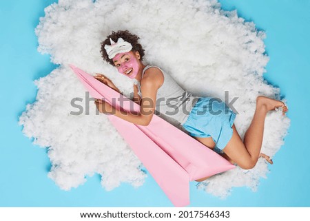 Joyful curly haired young woman in nightwear pretends flying has good fantasy holds big paper plane applies beauty mask on face poses on white cloud against blue background going to have sleep Royalty-Free Stock Photo #2017546343