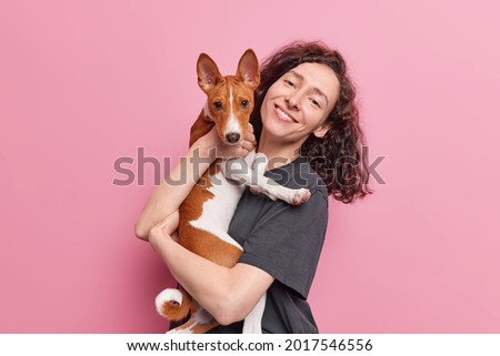 People and pets concept. Positive natural woman with curly dark hair carries her basenji dog spend time together have friendly relationship have walk pose against pink background. Happy pet owner Royalty-Free Stock Photo #2017546556