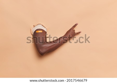 Photo of faceless dark skinned man makes rock n roll hand gesture breaks through beige paper background shows adhesive plaster on arm gets vaccination shot. Immunization and health care concept