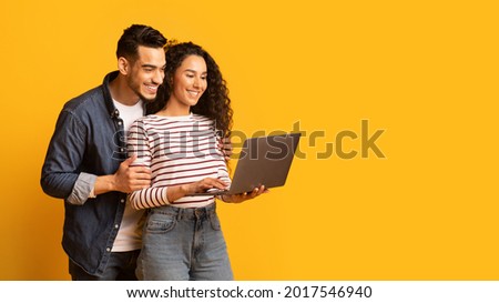 Young middle eastern couple using laptop together while standing over yellow background in studio, cheerful arab spouses shopping online or browsing new website on computer, panorama with copy space