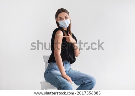Coronavirus vaccination saves lives. Young woman in face mask pointing at adhesive bandage on her arm, getting vaccinated against covid-19 on light background. Immunization for infectious disease Royalty-Free Stock Photo #2017556885