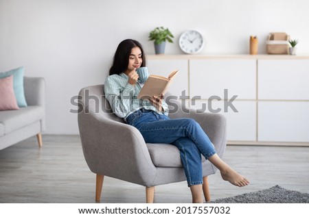 Attractive Indian lady with cup of aromatic coffee and open book relaxing in armchair at home. Pretty millennial woman enjoying hot beverage and reading exciting story, having lazy weekend indoors Royalty-Free Stock Photo #2017557032
