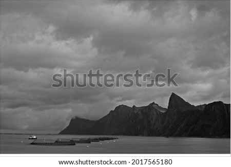 Black and white picture of boat, fish farm, mountains in the background in a fiord