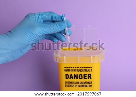 Doctor throwing used syringe needle into sharps container on violet background, closeup Royalty-Free Stock Photo #2017597067