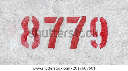 Red Number 8779 on the white wall. Spray paint. Number eight thousand seven hundred and seventy nine.