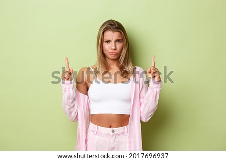 Image of gloomy, disappointed blond girl in pink outfit, complaining at something bad, pointing fingers up and frowning, standing over green background
