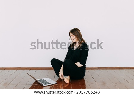 A freelance businesswoman is a young woman in casual clothes talking on the phone working using wireless technologies sitting on the floor against a white wall in an empty room at home