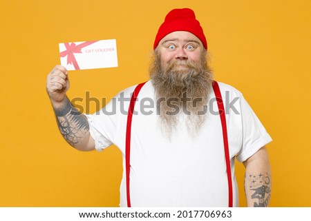 Fat pudge obese chubby overweight tattooed bearded surprised caucasian man 30s has big belly in white t-shirt red hat suspenders hold gift voucher flyer mock up isolated on yellow background studio