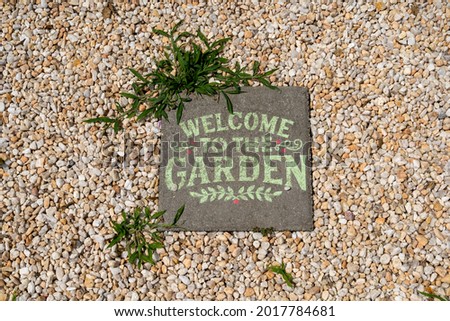 welcome to the garden sign in a gravel path with weeds growing beside it