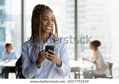 Dreamy happy young african american female manager employee worker holding smartphone in hands, looking in distance in modern workplace, thinking of new challenges or opportunities, copy space.