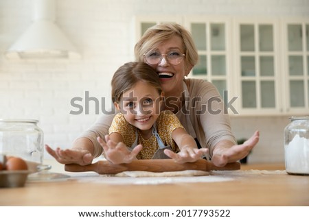 Head shot portrait happy mature grandmother in glasses with granddaughter rolling dough together, looking at camera, excited senior woman with adorable little girl cooking cookies in kitchen Royalty-Free Stock Photo #2017793522