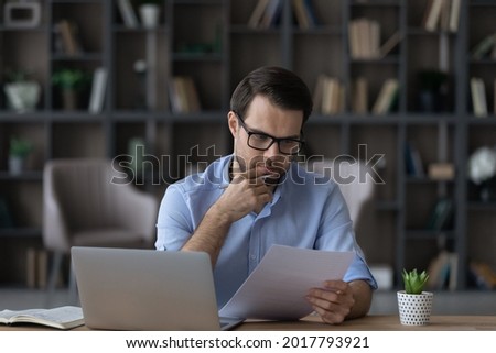 Working with papers. Serious millennial male entrepreneur work at home office alone hold read document hardcopy think on conditions terms. Focused young man engaged in paperwork edit text of agreement Royalty-Free Stock Photo #2017793921