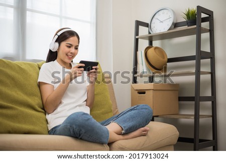 Beautiful young asian women play mobile game and put on wireless headphone sitting on sofa in living room. Playing game on smartphone winning victory moment. Very enjoy and fun relax time