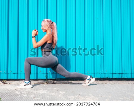 Fitness smiling woman in grey sports clothing with pink hair. Young beautiful model with perfect body.Female posing in the street near blue wall.Cheerful and happy. Stretching out before training