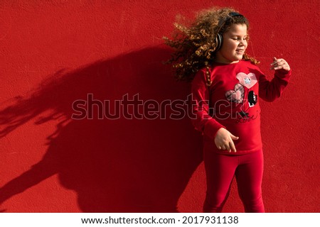 Little girl jumping and dancing enjoying the music with headphones. Red clothes and wall on background.