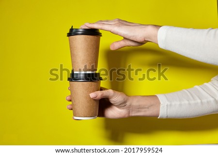 Hands holding two cups brown paper with black lid. Two coffee special offer or promo. Hands holding two cups on yellow screen background. Tea or Coffee to go. Brown paper cup with black lid.