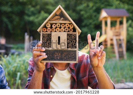Happy young man with bug hotel spending time outdoors in backyard, showing v sign with fingers.
