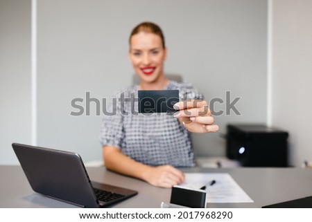 Young confident woman sitting in her office and looking at camera while giving business card. Plain gray wall in background.