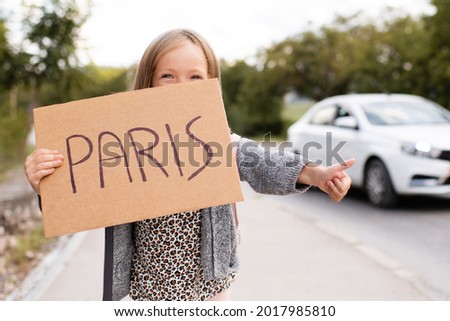 Happy smiling child girl 3-4 year old hold paper craft Paris handwritten sign hitch hiking on road outdoors. Happiness. Little kid toddler travel wear casual clothes and backpack. Summer vacation time Royalty-Free Stock Photo #2017985810