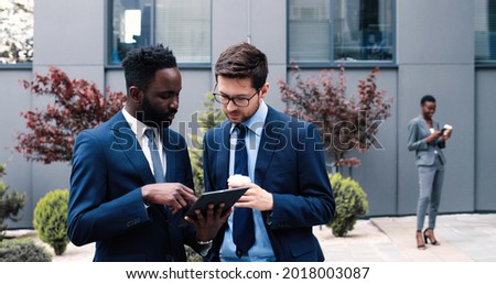 Confident business. Waist-up portrait of young serious businessman is standing with tablet near his multiracial male colleague while looking at the screen intently. Business concept 