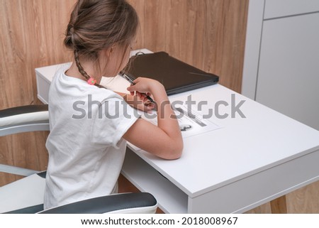 A little girl studies at home at a white table performs an educational task with a pen in her hands. A black laptop lies on a white table