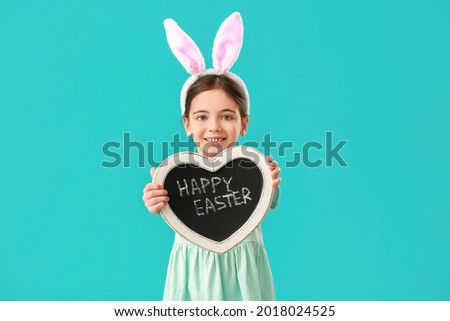 Cute little girl with bunny ears holding chalkboard with text HAPPY EASTER on color background