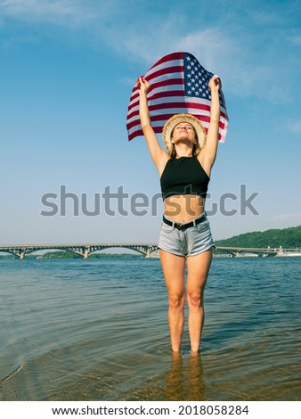 A young woman in denim shorts with the US flag stands on a river in the water against the background of a long bridge in the distance. Independence Day. Celebration and freedom
