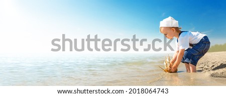 Kid play on the beach on a hot sunny day. Little girl dressed as a sailor stands barefoot on the sandy shore and launches a boat into the sea. Child dreams of travel and adventure. Royalty-Free Stock Photo #2018064743