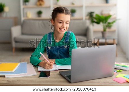 Online Education Concept. Smiling young teenage girl sitting at desk, using looking at laptop screen writing in her notebook. Schoolgirl watching online course, taking notes, doing homework Royalty-Free Stock Photo #2018070413