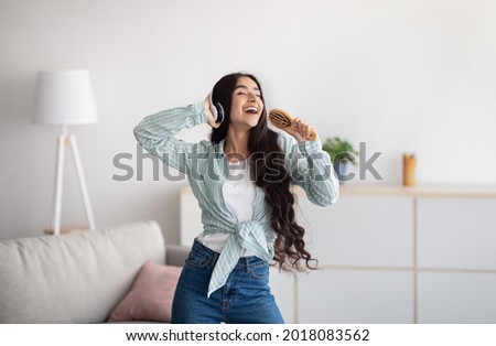 Beautiful Indian lady with headphones dancing and singing, using hairbrush as microphone at home. Millennial woman moving to her favorite song, enjoying music, pretending to be popular star Royalty-Free Stock Photo #2018083562