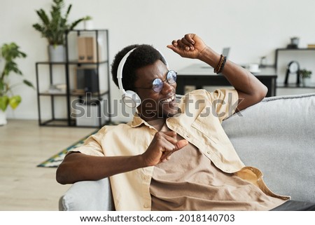 African happy man in wireless headphones sitting on the sofa dancing and enjoying the music at home
