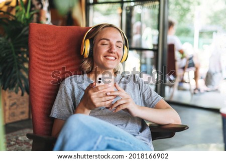Young smiling blonde woman with close eyes in yellow headphones enjoys music with mobile phone sitting on chair at the cafe Royalty-Free Stock Photo #2018166902