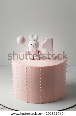 Birthday cake with pink cream cheese frosting decorated with rabbit holding balloon made of mastic. Little baby girl one year old celebrating. First birthday party